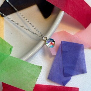 May include: A silver chain necklace with a round pendant featuring a colorful abstract design. The pendant is made of glass and has a clear, shiny finish.