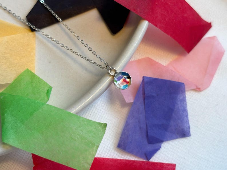 May include: A silver chain necklace with a round pendant featuring a colorful abstract design. The pendant is made of glass and has a clear, shiny finish.