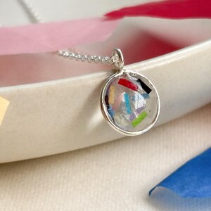 Puede incluir: Un collar con colgante de plata con un colgante redondo de resina transparente lleno de piezas coloridas tipo confeti. El colgante cuelga de una delicada cadena de plata. El collar se exhibe sobre un plato de cerámica blanca con detalles de papel de colores.