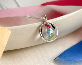 May include: A silver pendant necklace with a round, clear resin pendant filled with colorful, confetti-like pieces. The pendant hangs from a delicate silver chain. The necklace is displayed on a white ceramic dish with colorful paper accents.