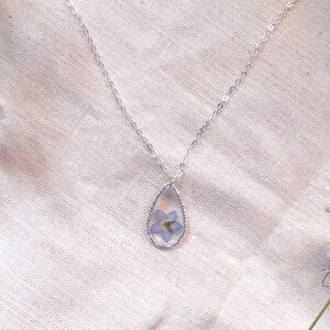 May include: A silver chain necklace with a teardrop-shaped pendant. The pendant contains a pressed blue flower.