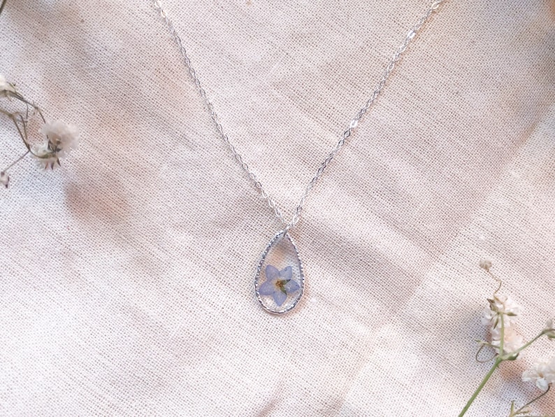 May include: A silver chain necklace with a teardrop-shaped pendant. The pendant contains a pressed blue flower.