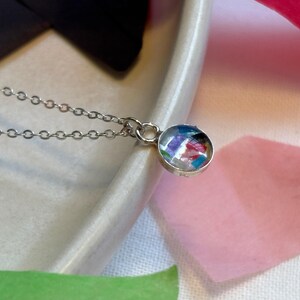 May include: A silver chain necklace with a round pendant featuring a colorful, abstract design. The pendant is made of clear resin with embedded pieces of paper.