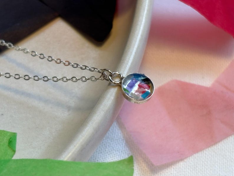 May include: A silver chain necklace with a round pendant featuring a colorful, abstract design. The pendant is made of clear resin with embedded pieces of paper.