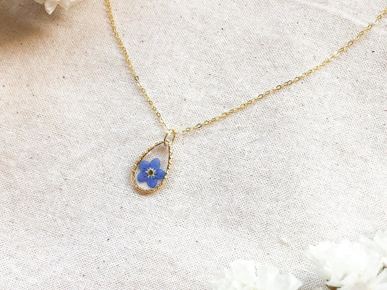 May include: A delicate gold chain necklace with a teardrop-shaped pendant featuring a pressed blue forget-me-not flower.