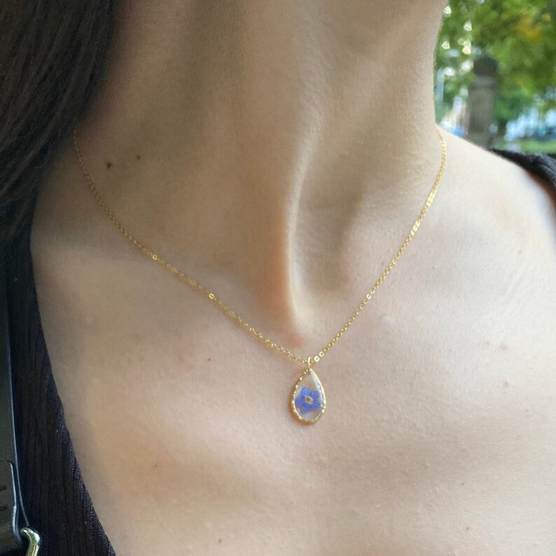May include: A gold chain necklace with a teardrop-shaped pendant featuring a pressed blue flower.