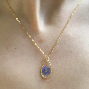 May include: A gold chain necklace with a teardrop-shaped pendant featuring a preserved blue flower.