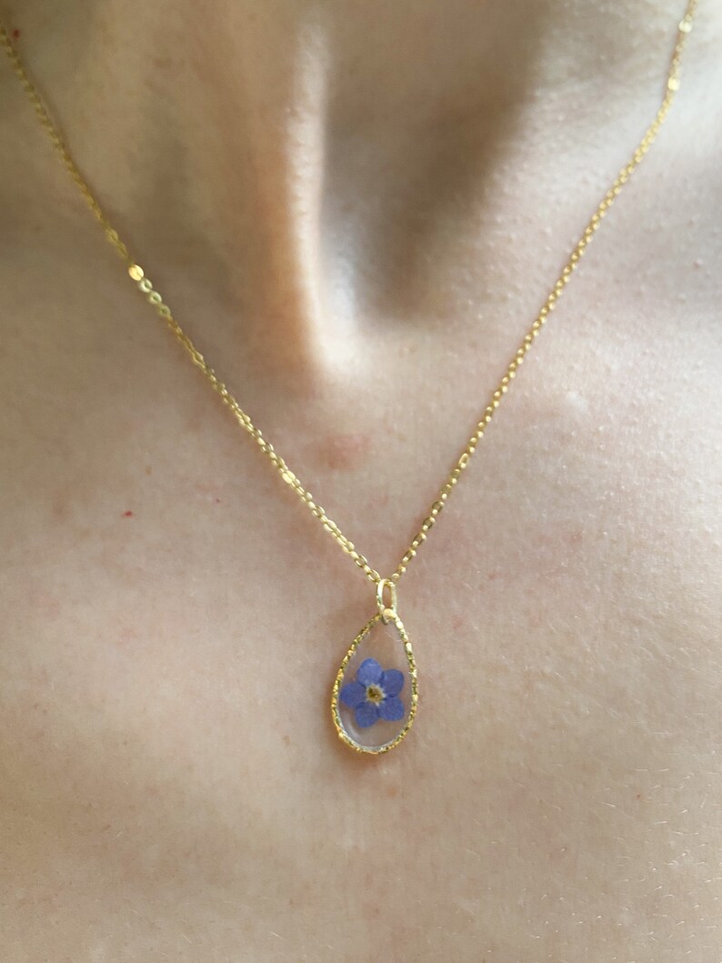 May include: A gold chain necklace with a teardrop-shaped pendant featuring a preserved blue flower.