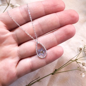 May include: A silver chain necklace with a teardrop-shaped pendant featuring a pressed purple flower encased in clear resin.
