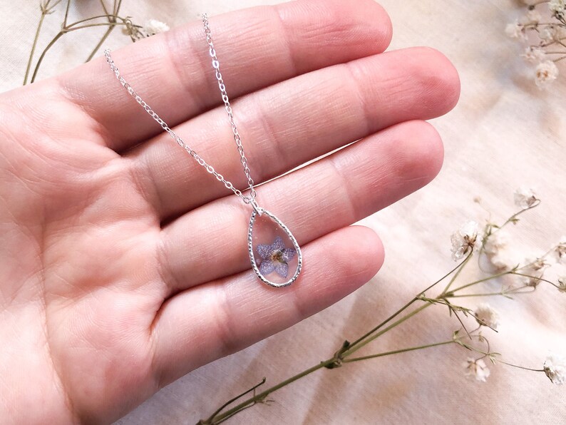 May include: A silver chain necklace with a teardrop-shaped pendant featuring a pressed purple flower encased in clear resin.