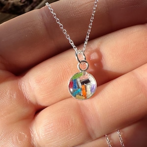 May include: A silver necklace with a small, round pendant. The pendant is filled with colorful, abstract shapes in shades of blue, orange, green, and purple. The chain is delicate and appears to be made of silver.