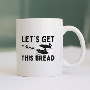 Può includere: Una tazza da caffè in ceramica bianca con una silhouette nera di anatre e il testo "Let's get this bread".
