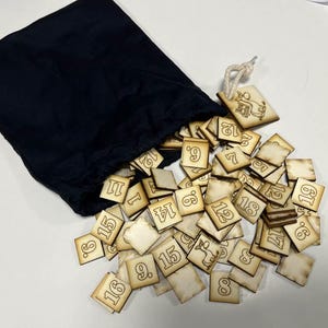 Op de afbeelding: Een zwarte trekkoordtas met een stapel houten tegels die eruit vallen. De tegels zijn genummerd van 1 tot 18 en bevinden zich in verschillende posities.