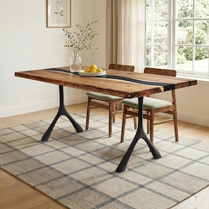 May include: A rectangular wooden dining table with a black resin river design, supported by black metal legs. Two wooden chairs with green cushions are positioned behind the table. The table is set on a patterned rug. A vase with branches and a bowl of lemons are on the table.