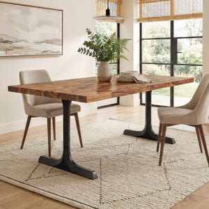 May include: A rectangular wooden dining table with a dark finish, supported by black metal legs. Two beige upholstered chairs are positioned on either side. A vase of greenery and a stack of plates sit on the table. The rug is beige with a geometric pattern.