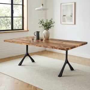 May include: A rectangular wooden dining table with a natural wood grain finish. The table is supported by two black, Y-shaped metal legs. A white vase with greenery, a gray pitcher, and two glasses are on the table. The table is set on a light-colored rug.