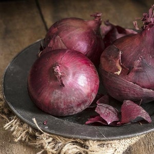 May include: Three red onions on a dark gray plate. The onions are whole and have their skins on. The plate is on a burlap surface.