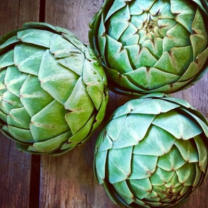 May include: Three fresh artichokes with green and light green leaves are arranged on a wooden surface. The artichokes have a layered, textured appearance, with each leaf overlapping the next. The wooden surface has a warm, brown tone.