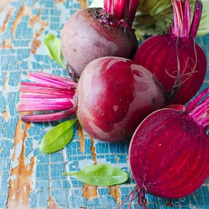 May include: Four red beets, two whole and two halved, with green leafy tops, on a blue and brown textured surface.