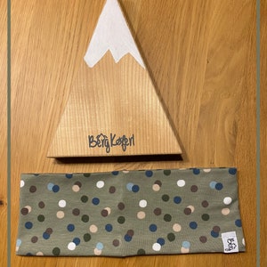 Könnte beinhalten: Ein grünes Stirnband mit weißen, braunen und blauen Punkten. Das Stirnband wird auf einem hölzernen, bergförmigen Ständer mit dem Text "Berg Koferl" auf der Vorderseite präsentiert.