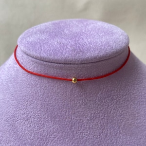 May include: A red cord necklace featuring a small, gold-coloured bead at its centre. The necklace is displayed on a purple velvet bust. The cord is thin, and the bead is smooth and round.