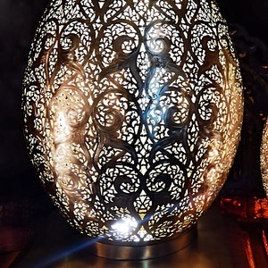 Peut inclure: Lampe en métal doré en forme d'œuf, ornée d'un motif ajouré complexe. Elle repose sur un socle rectangulaire assorti, également ajouré. Le design permet à la lumière de filtrer, créant une ambiance chaleureuse.