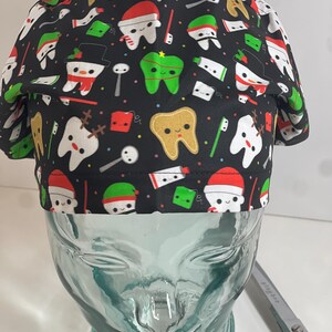 May include: A black scrub hat with a Christmas-themed print featuring cartoon teeth, toothbrushes, toothpaste, and other dental-related items. The print also includes holiday elements like snowmen, Santa hats, and candy canes.