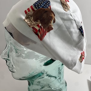 May include: White surgical scrub cap featuring a brown dog's head and American flag design. The cap is made of a soft, lightweight fabric and is designed to fit comfortably. The patriotic theme makes it a unique accessory.