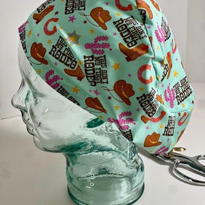 May include: A light teal scrub cap with a rodeo theme. The cap features a repeating pattern of cowboy boots, hats, horseshoes, and cacti in brown, pink, and yellow. The text "This ain't my first rodeo" is printed on the cap.