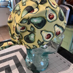 May include: A green and yellow fabric scrub hat with a pattern of avocados. The hat is on a glass head form.