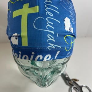 May include: Blue surgical scrub hat with a white and green print featuring a cross, sheep, and the words "Hallelujah", "Rejoice", and "He is Risen!"