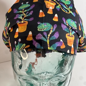 May include: A black scrub cap featuring a whimsical pattern of green frog-like creatures with purple and teal plant-like bodies, orange pots, and candy corn. The cap is made of a soft fabric and is designed to fit comfortably.