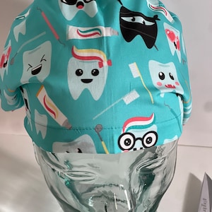 May include: A turquoise blue scrub hat with a fun dental themed print. The print features cartoon teeth with different expressions, toothbrushes, and toothpaste tubes.