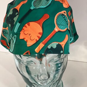 May include: Green scrub hat with a repeating pattern of orange cats and teal tennis rackets.