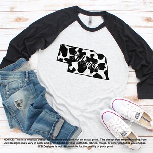 May include: A raglan baseball tee with a gray body and black sleeves. The shirt features a black and white cow print outline of Nebraska with the word "Nebraska" in script. Blue jeans and white sneakers are also in the image.