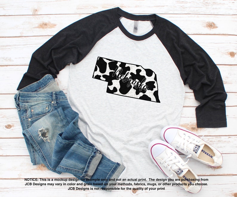 May include: A raglan baseball tee with a gray body and black sleeves. The shirt features a black and white cow print outline of Nebraska with the word "Nebraska" in script. Blue jeans and white sneakers are also in the image.