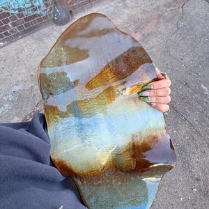May include: A handcrafted, organic-shaped ceramic platter with a glossy finish. The color palette includes shades of brown, blue, and gold, creating a unique, marbled effect. The platter's irregular edges add to its artistic appeal.