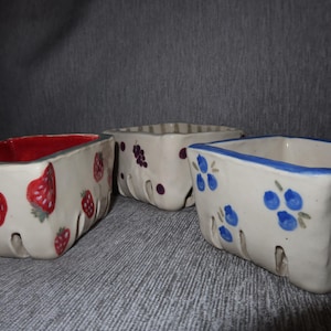 May include: Three ceramic berry baskets. One has red strawberries, another has purple grapes, and the third has blue blueberries. Each basket is white with a different fruit design and a different colored rim.