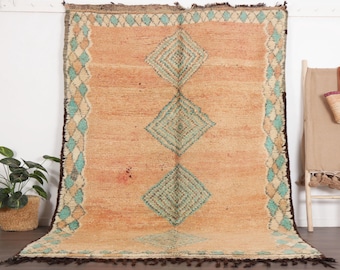 May include: A rectangular area rug featuring a warm peach-coloured field with three large, centred diamond motifs in teal and cream. The border consists of a repeating diamond pattern in teal and cream, finished with dark brown fringe at both ends.