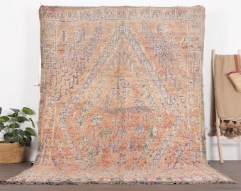 May include: A large, rectangular, vintage rug with a peach and blue geometric design. The rug is displayed against a white wall, with a wooden ladder and a woven basket in the background. The rug's pattern includes diamond and linear shapes.