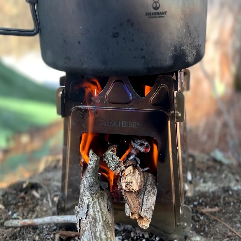Peut inclure: Un r&eacute;chaud de camping en m&eacute;tal noir avec une casserole sur le dessus. Le r&eacute;chaud est allum&eacute; et des flammes sortent du bas. Le r&eacute;chaud est marqu&eacute; "Coopers Bay."