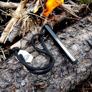 May include: A black fire starter rod with a black cord and a white tag with a logo. The fire starter is on a log with a fire burning in the background.