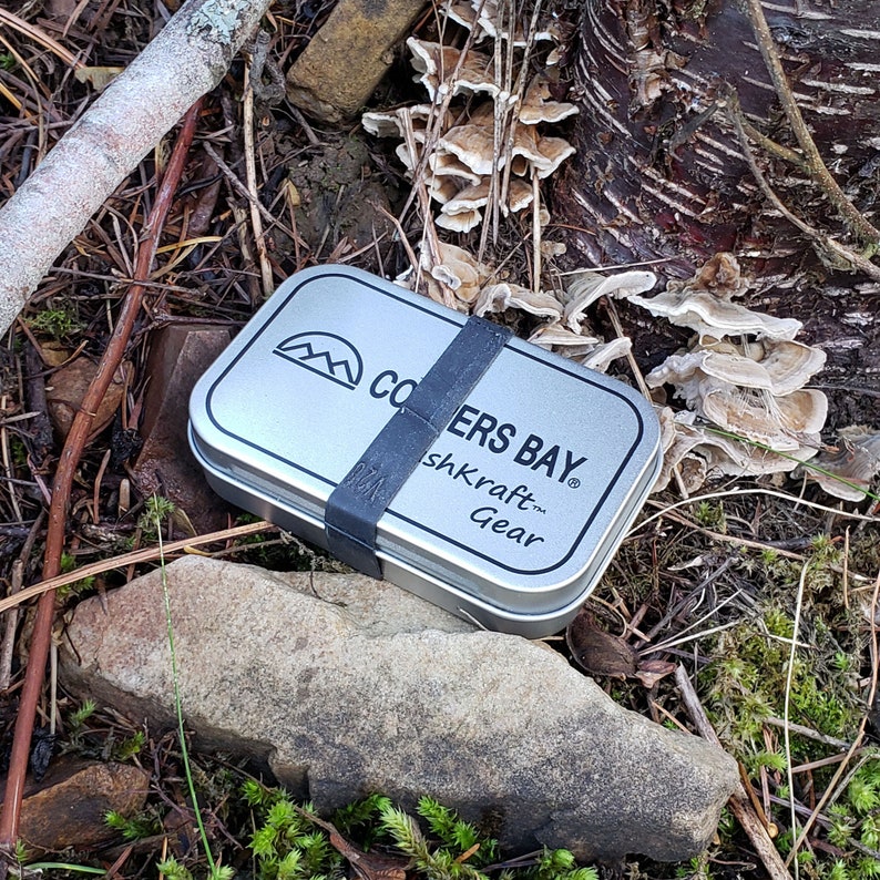 Puede incluir: Una lata de metal plateada con una banda de goma negra alrededor. La lata tiene las palabras "Coopers Bay" y "shKraft Gear" impresas en ella. La lata est&aacute; sentada en un lecho de hojas marrones y hongos.