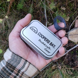 Puede incluir: Una lata de plata con el texto "Coopers Bay&reg; Bushkraft&trade; Gear" en ella. La lata est&aacute; siendo sostenida en una mano, y un anillo de goma negro est&aacute; en un dedo de la misma mano.