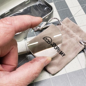 Puede incluir: Una lupa redonda con un logotipo negro que dice "Coops Bay" y un gr&aacute;fico de cordillera. La lupa est&aacute; sobre una bolsa de gamuza marr&oacute;n claro.