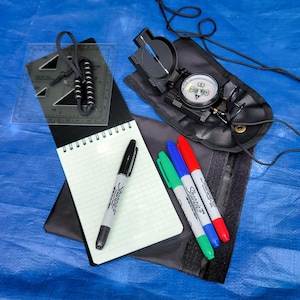 May include: A black folding compass with a lanyard, a clear plastic protractor with a beaded cord, a spiral-bound grid notebook, and three Sharpie markers in black, blue, and red. The items are arranged on a black fabric case.