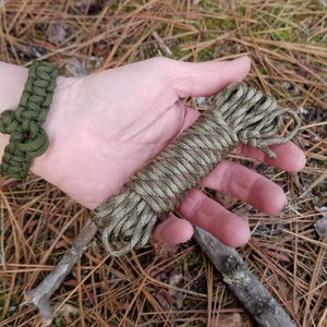 May include: A close-up of a hand holding a green and brown camouflage paracord. The paracord is coiled and ready to be used for crafting or survival purposes.