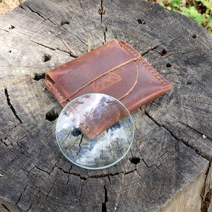 May include: A brown leather wallet with a magnifying glass on top of it. The magnifying glass is reflecting the sky and trees.