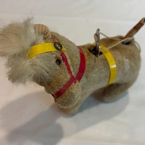May include: A vintage wind-up toy horse covered in tan fuzzy material with a cream-colored mane. The horse features a red harness, yellow straps, and a metal winding key attached to its back. It has dark eyes and a small tail.
