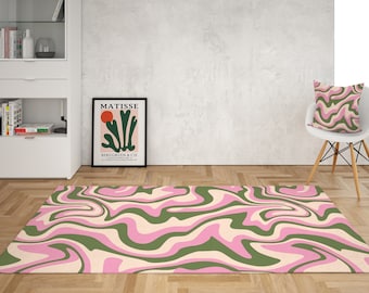 May include: A pink and green abstract rug with a swirling pattern. The rug is on a light wood floor in a room with a white wall and a white chair with a pillow.